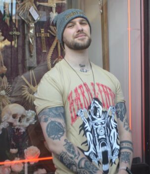 A photo of Iain, who has a beard and several tattoos. He is wearing a yellow tshirt and a grey beanie that say 'Hard Lines'.